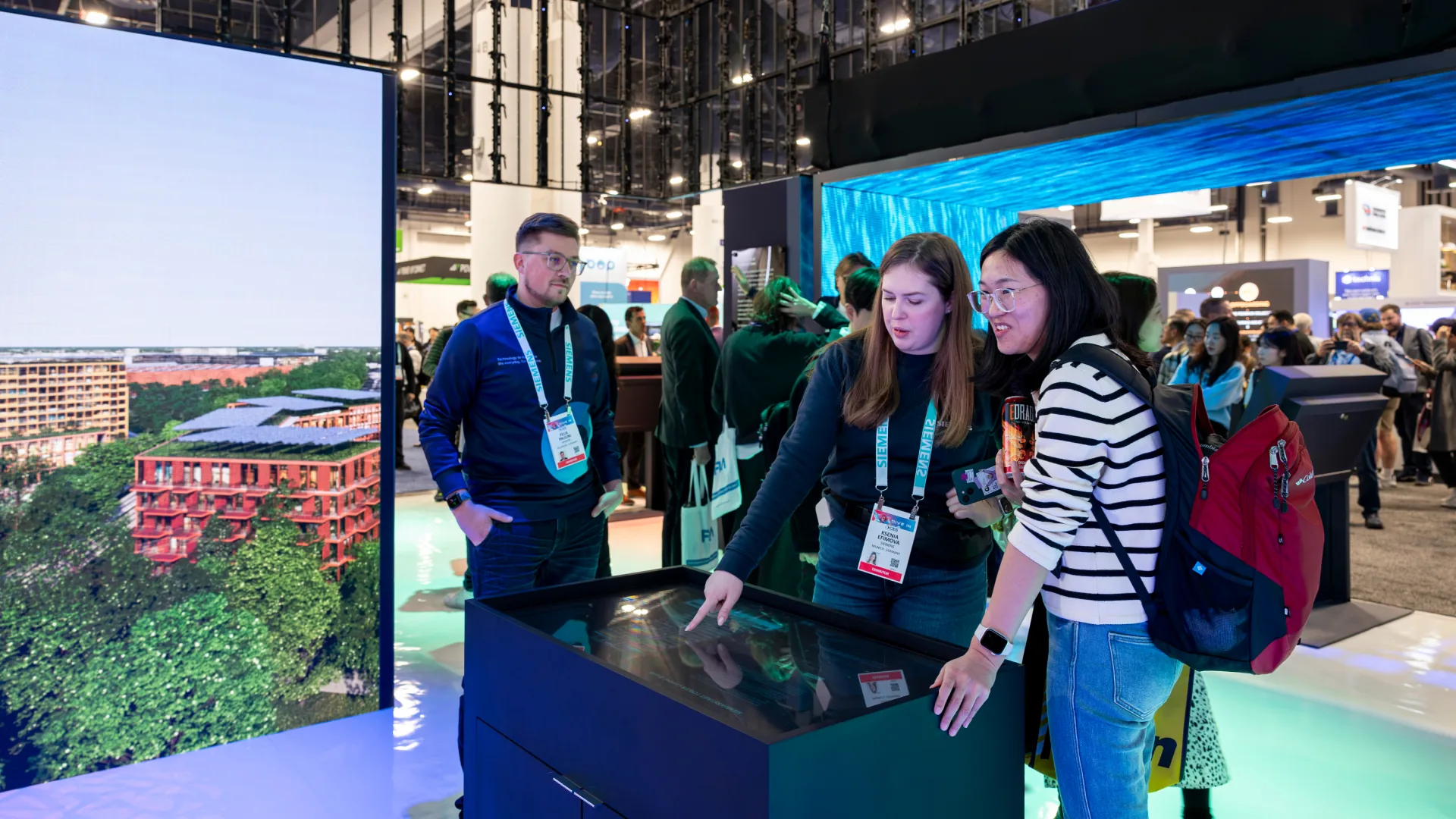 Two attendees explore a touch table beside a screen showing a red-roofed building