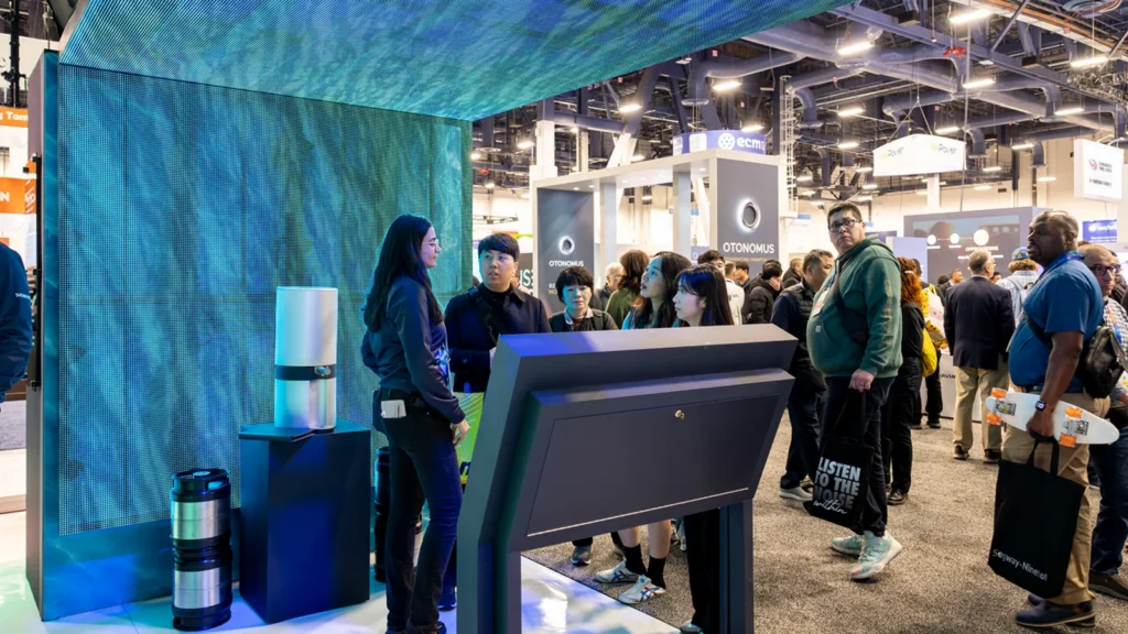 Siemens booth at CES 2025 featuring a Sparks-designed immersive LED tunnel — LED walls wrapping overhead as a ceiling canopy with ocean-wave graphics, a cylindrical product display, and attendees engaging with a Siemens host.