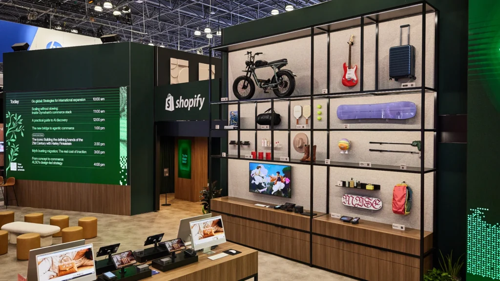 The Sparks-designed and built two-level Shopify booth at NRF 2026 at the Javits Center in New York, featuring the 16-foot curated merchant showcase wall with real merchant products (bike, guitar, luggage, yoga mat) alongside the workshop schedule LED and checkout demo counter.