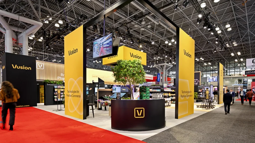 The Vusion booth at NRF 2026 with bold yellow branding and the 'Technologies for Positive Commerce' tagline, a clean minimal architecture that reinforced the show's shift toward clarity and restraint on the NRF Big Show floor at the Javits Center in New York.