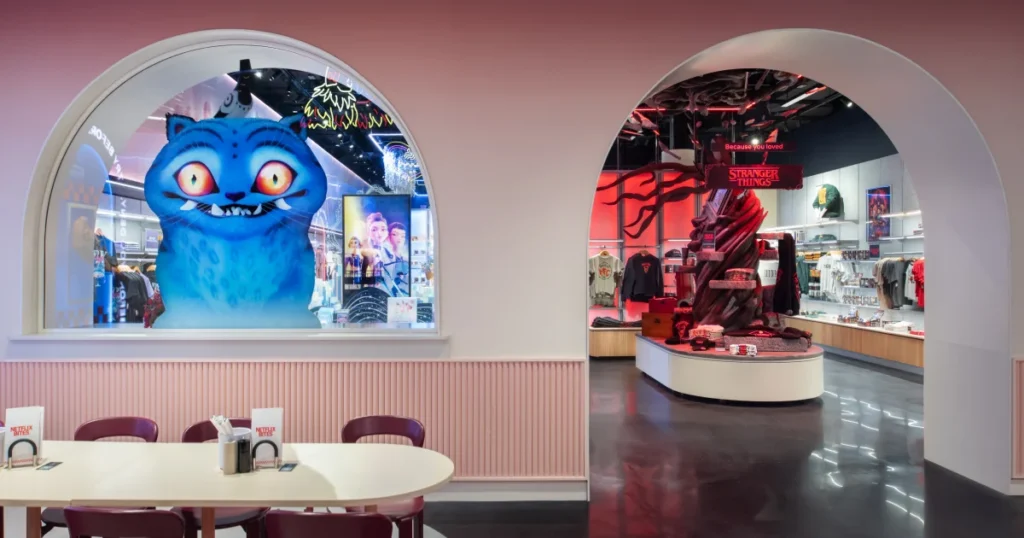 Pastel pink interior of Netflix House Philadelphia featuring arched window vignettes for Wednesday and Stranger Things title worlds, Netflix Bites cafe seating in the foreground.