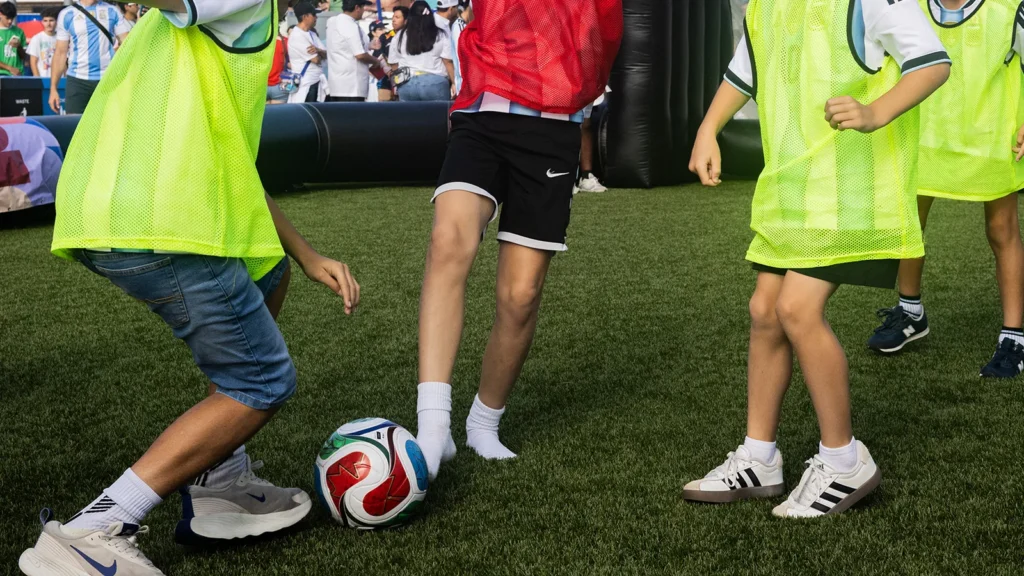 Youth soccer players in scrimmage vests around a FIFA-branded ball on turf at a Sparks-produced FIFA World Cup 2026 fan experience — close-up moment of play with Argentina jersey supporters in the background.