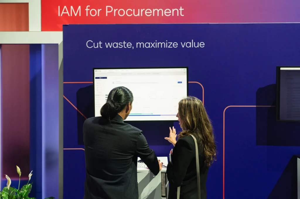 IAM for Procurement demonstration—"Cut waste, maximize value"—with attendees reviewing the platform at Momentum 2026.