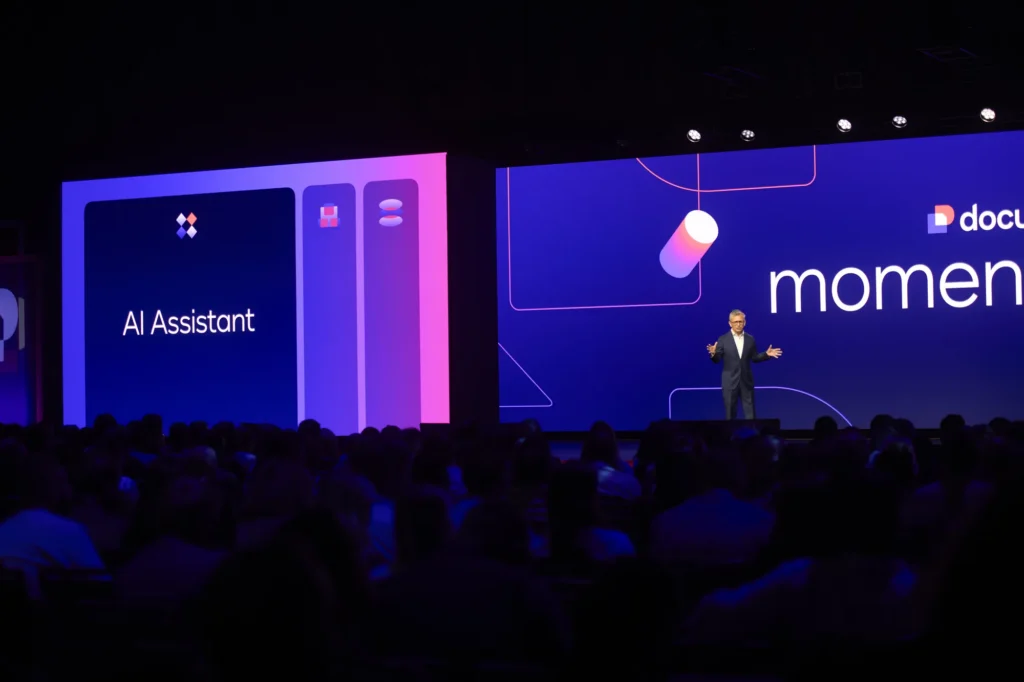 Docusign Momentum 2026 keynote showing the IAM AI Assistant on a large-format LED stage above a seated audience.