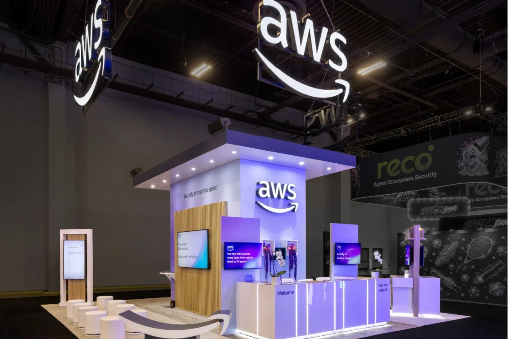 Sparks-designed and built AWS 30x30 exhibit at Black Hat 2026 with dual illuminated AWS logos suspended overhead, warm wood-slat walls, and "Security at machine speed" messaging on displays at Mandalay Bay Convention Center in Las Vegas.
