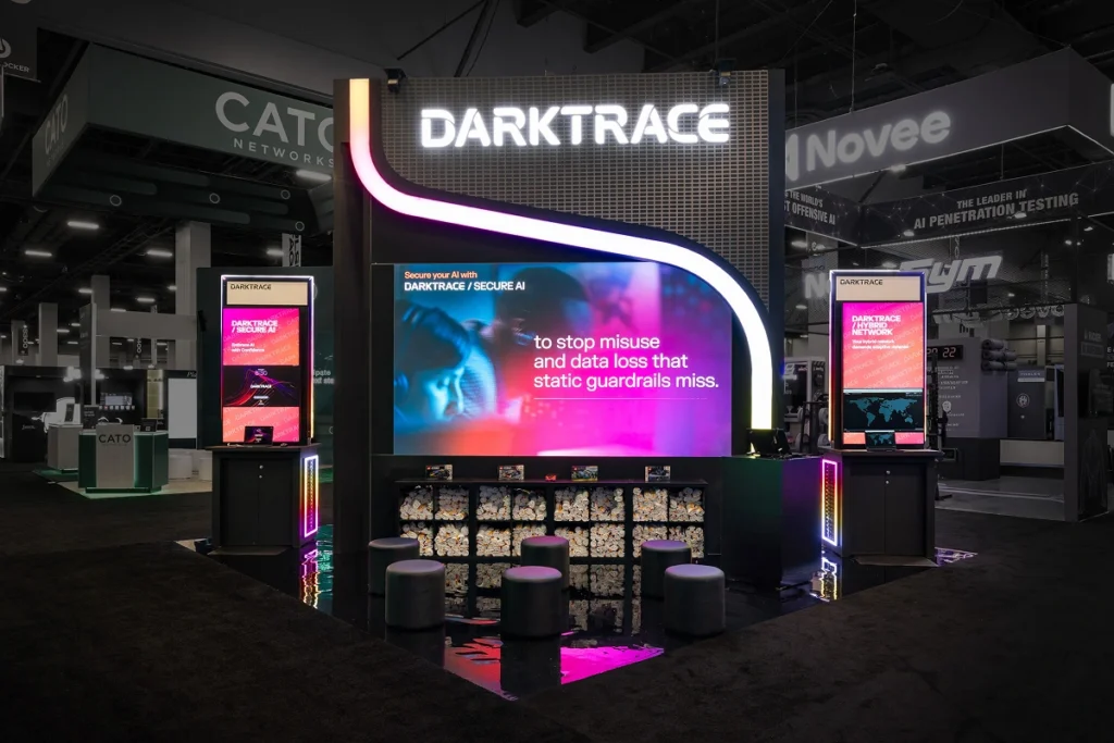 Sparks-designed and built Darktrace 20x20 exhibit at Black Hat 2026 with an illuminated DARKTRACE lightbox marquee, curving magenta LED accent, and "Darktrace / Secure AI" messaging at Mandalay Bay Convention Center in Las Vegas.