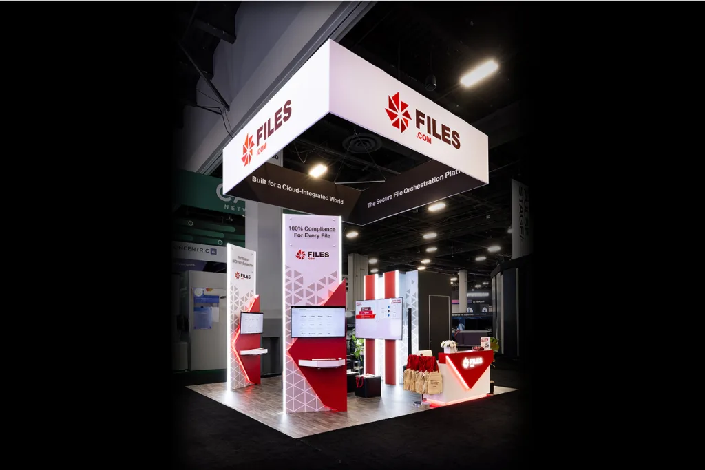 Sparks-designed and built Files.com 20x20 exhibit at Black Hat 2026 with an illuminated FILES.COM overhead sign, red-and-white geometric branding, "100% Compliance For Every File" messaging, and product demo stations at Mandalay Bay Convention Center in Las Vegas.