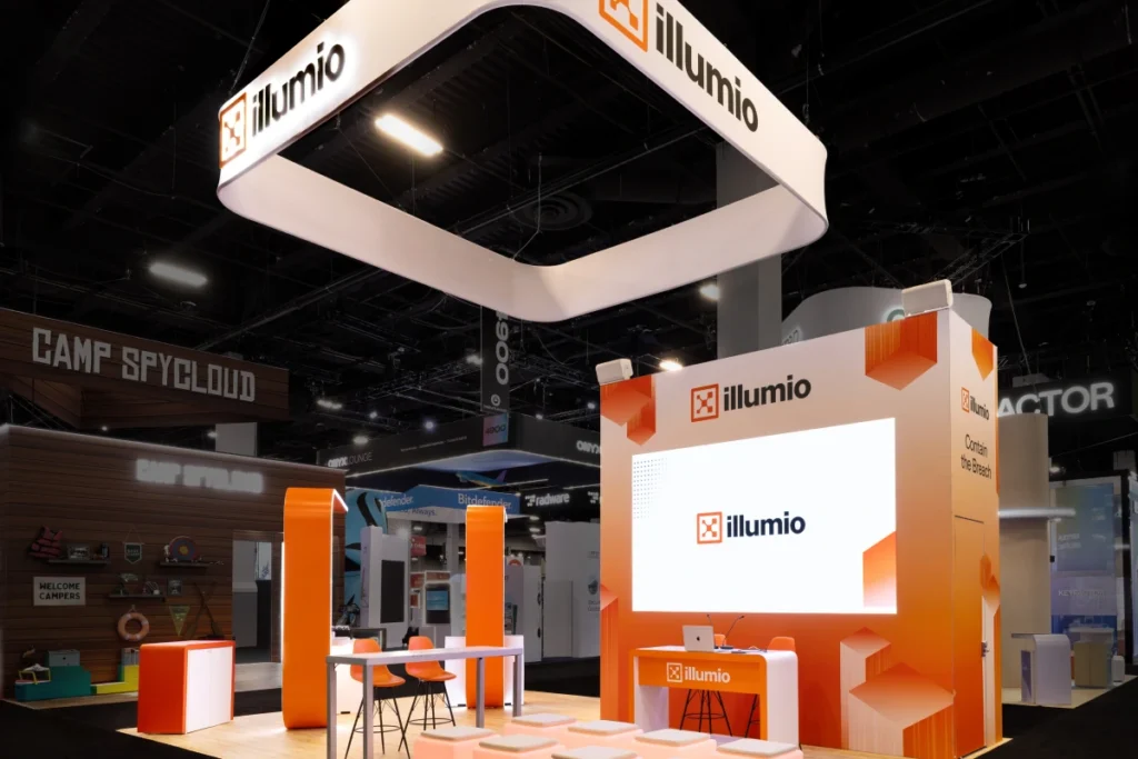 Sparks-designed and built Illumio 20x30 exhibit at Black Hat 2026 with a suspended square canopy carrying the illumio logo, orange corner columns, illuminated cube stools, and a "Contain the Breach" video wall at Mandalay Bay Convention Center in Las Vegas.