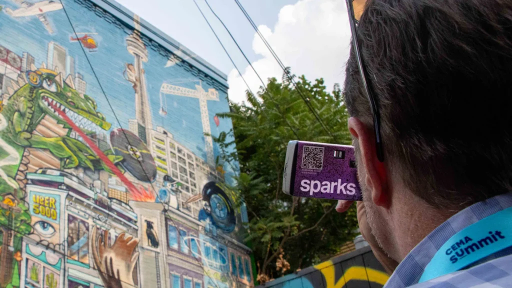 A CEMA Summit 2026 attendee — CEMA Summit lanyard visible — photographs a massive Toronto Graffiti Alley mural featuring the CN Tower, a cartoon alligator, and Uber 5000 street art with a purple Sparks-branded disposable camera during the Sparks-led walking tour of Toronto's Graffiti Alley.