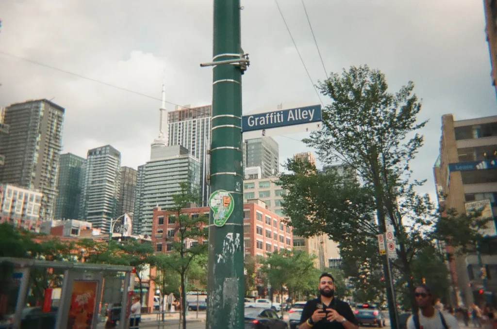 Sparks-hosted CEMA Summit 2026 disposable-camera photo — a Graffiti Alley street sign in downtown Toronto with the city skyline in the background, captured by a CEMA Summit attendee on the Sparks-led Graffiti Alley walking tour.