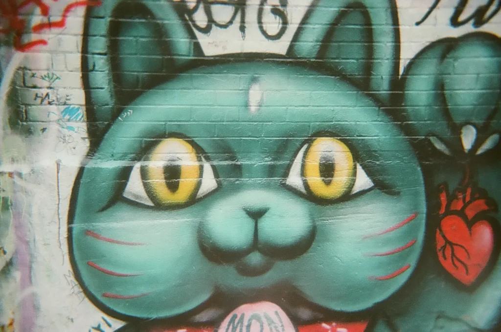 Sparks-hosted CEMA Summit 2026 disposable-camera photo — a large green cat mural with big yellow eyes and a red heart on its chest in Toronto's Graffiti Alley, captured by a CEMA Summit attendee on the Sparks-led walking tour.