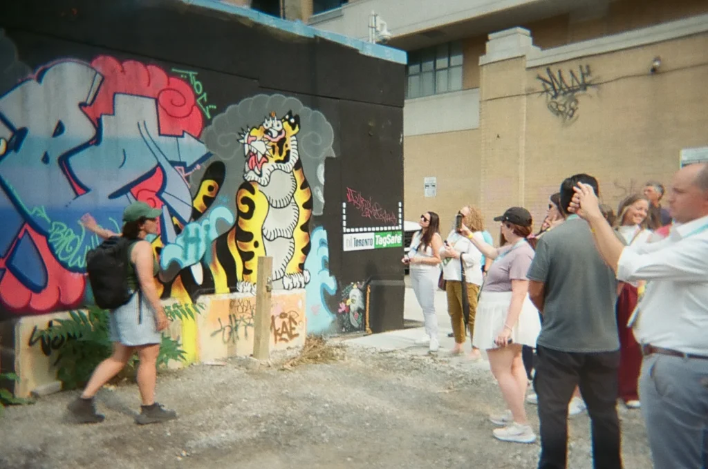 Sparks-hosted CEMA Summit 2026 disposable-camera photo — CEMA Summit attendees on the Sparks-led Graffiti Alley walking tour in Toronto, one photographing the murals with a disposable camera.