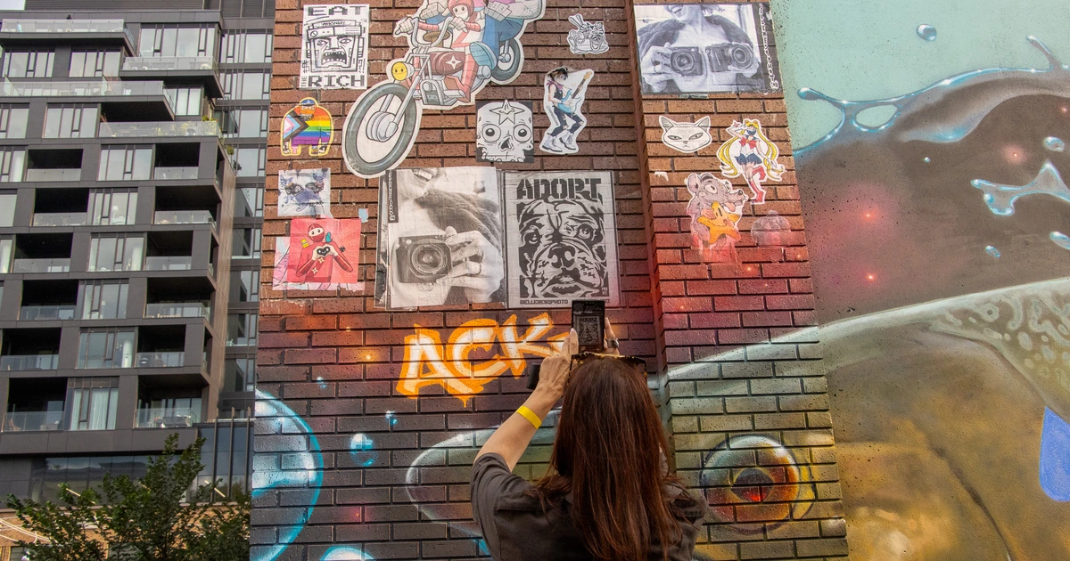 A Sparks-hosted CEMA Summit 2026 attendee photographs a Toronto street-art paste-up wall — with "Eat the Rich" and "Adopt" posters over vibrant tag murals — during the Sparks-led walking tour of Toronto's Graffiti Alley.