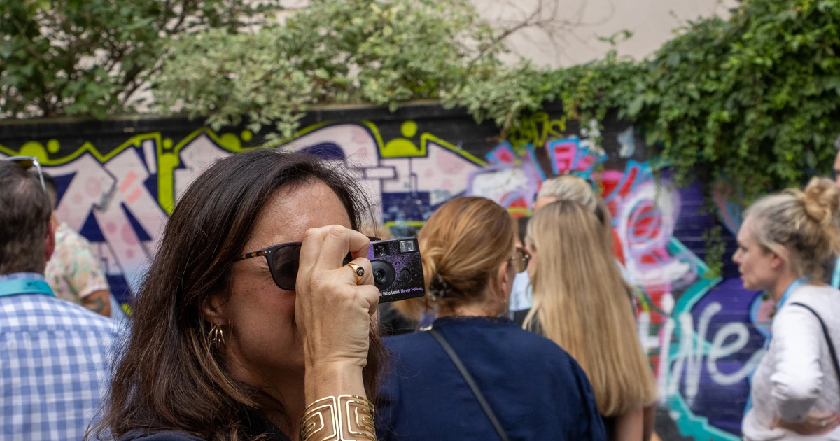 A Sparks-hosted CEMA Summit 2026 attendee shoots a graffiti mural with a Sparks-branded disposable camera printed with the Sparks tagline "Those Who Lead, Never Follow" during the Sparks-led walking tour of Toronto's Graffiti Alley.