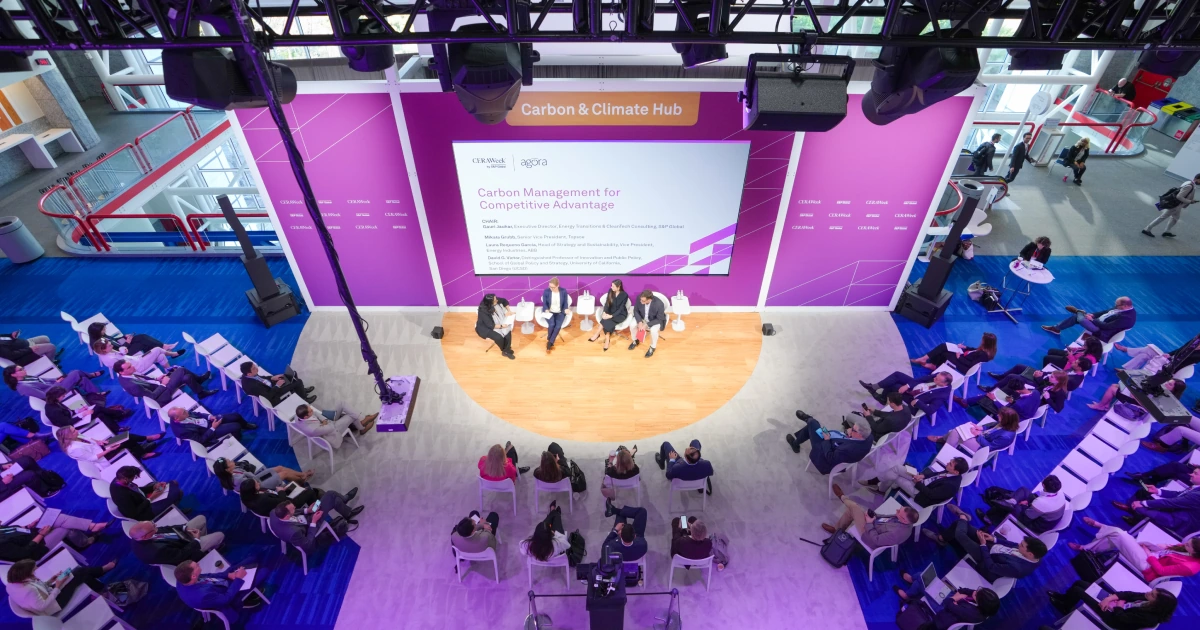 Overhead view of the Sparks-produced Carbon & Climate Hub in the Innovation Agora at CERAWeek 2026 by S&P Global — a four-person 'Carbon Management for Competitive Advantage' panel session in front of a seated audience under overhead lighting truss at the George R. Brown Convention Center in Houston.