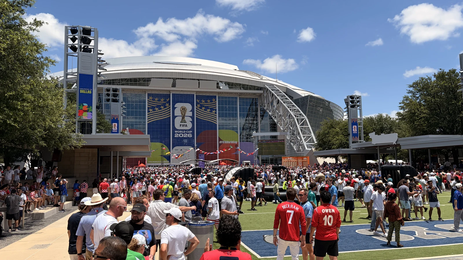 A massive fan crowd fills the Sparks-produced Stadium Fan Experience Town Square outside AT&T Stadium — the FIFA World Cup 2026 backdrop and main stage rise above the plaza at the Dallas host city, with England fans in Beckham 7 and Owen 10 jerseys in the foreground.