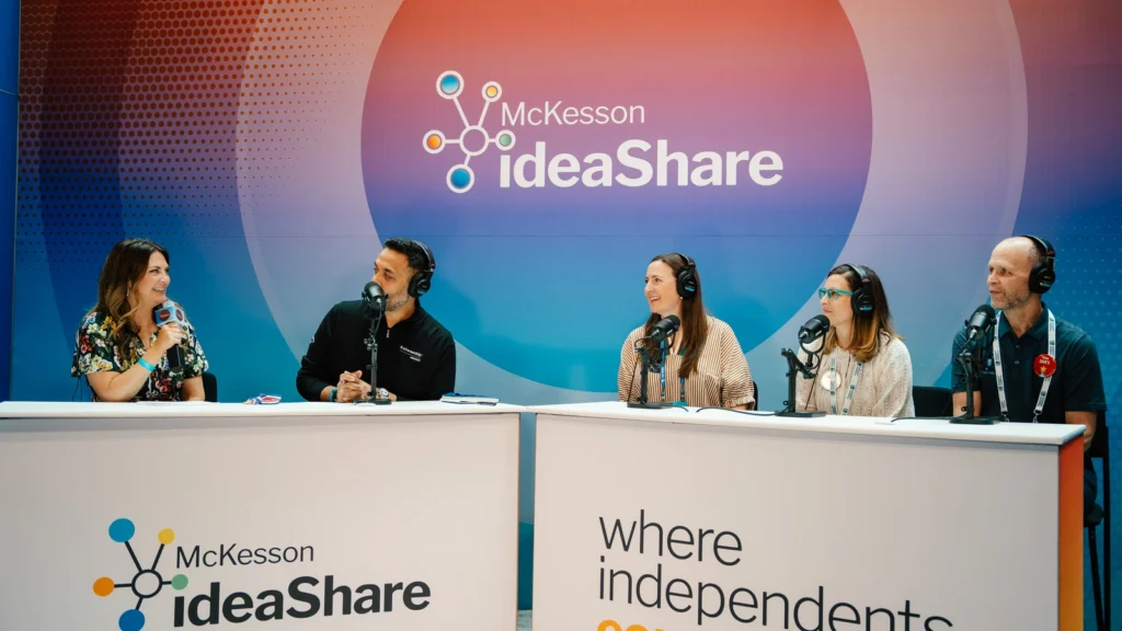 Five hosts and guests record a live podcast at the Sparks-designed McKesson ideaShare 2025 Broadcast Center — a brick storefront-style set with 'Where Independents Connect' tagline — in the Gaylord Opryland atrium outside the exhibit hall in Nashville.