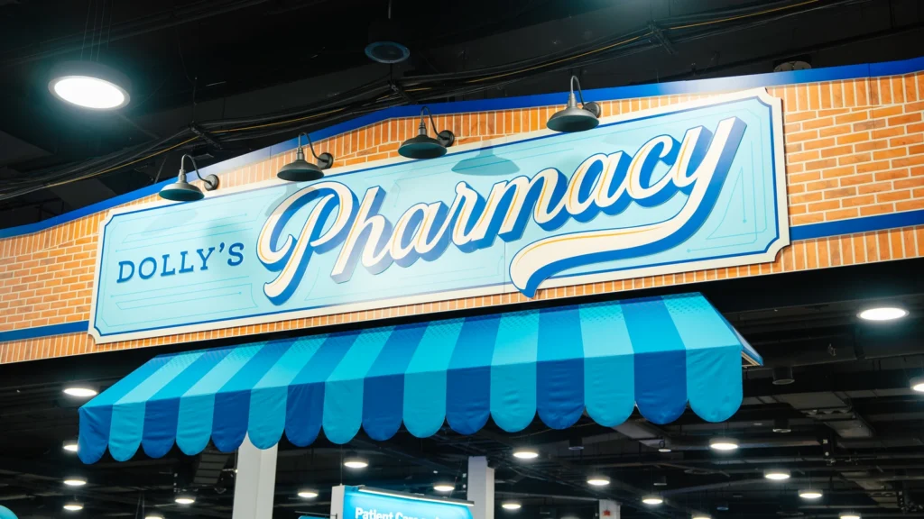 Close-up of the Sparks-designed Dolly's Pharmacy storefront — a brick facade with a blue-and-white striped awning and hand-lettered 'Ask us — we can help!' signage — on the Main Street USA-themed McKesson ideaShare 2025 tradeshow floor at the Gaylord Opryland Resort & Convention Center in Nashville.