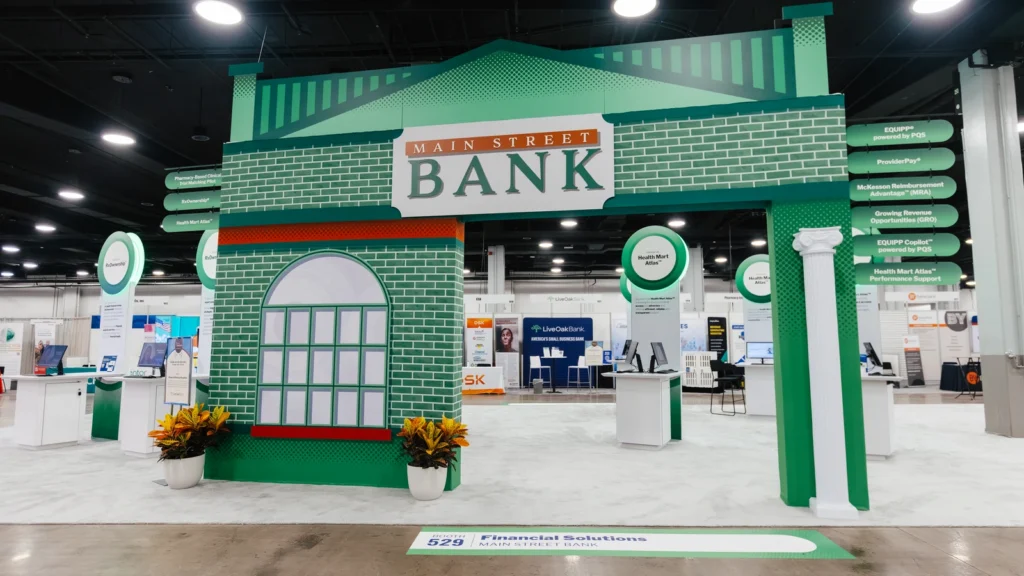 The Sparks-designed Main Street Bank storefront — a green-brick facade with white columns — on the Main Street USA-themed McKesson ideaShare 2025 tradeshow floor at the Gaylord Opryland Resort & Convention Center in Nashville.