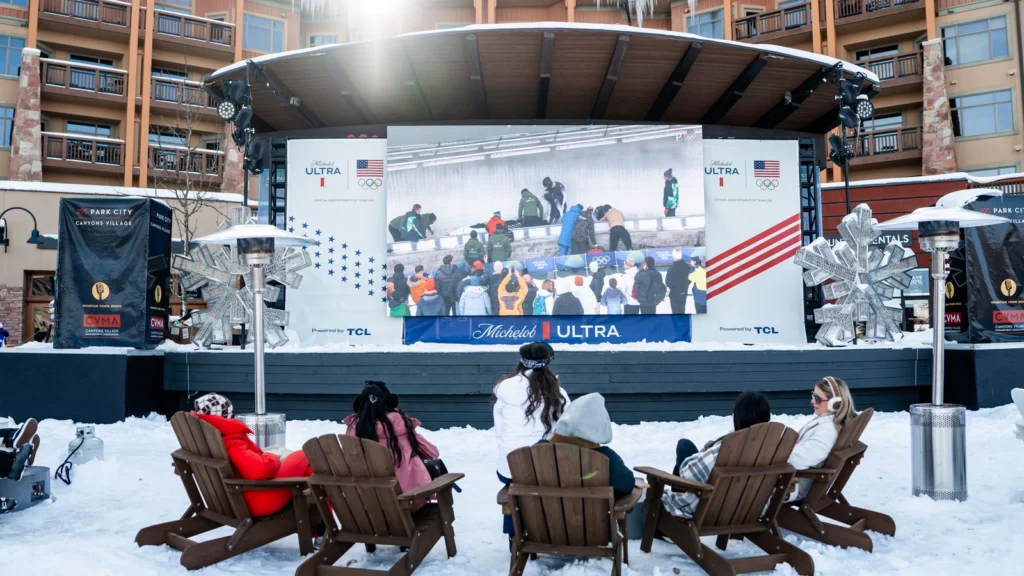 Adirondack watch-party seating in front of the Michelob ULTRA and Team USA main stage at Sparks-produced Base ULTRA Village, Canyons Village at Park City — TCL-powered Olympic coverage and outdoor Team USA celebration during the Milano Cortina 2026 Winter Olympics closing weekend.