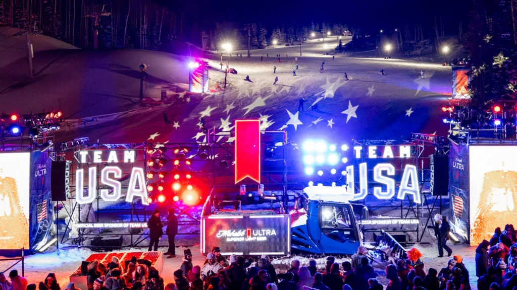 Sparks-designed night ski run at Michelob ULTRA Summit, Canyons Village at Park City — American flag stars projected onto the slope with Team USA light installations flanking the Michelob ULTRA snowcat, closing weekend of the Milano Cortina 2026 Winter Olympics.