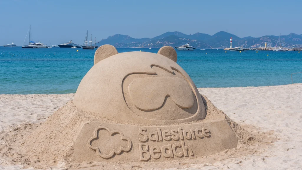 Salesforce Astro mascot sand sculpture with Salesforce Beach carved base, yachts and mountains on the Mediterranean horizon at the Sparks-designed Cannes Lions 2026 activation on Annex Beach.
