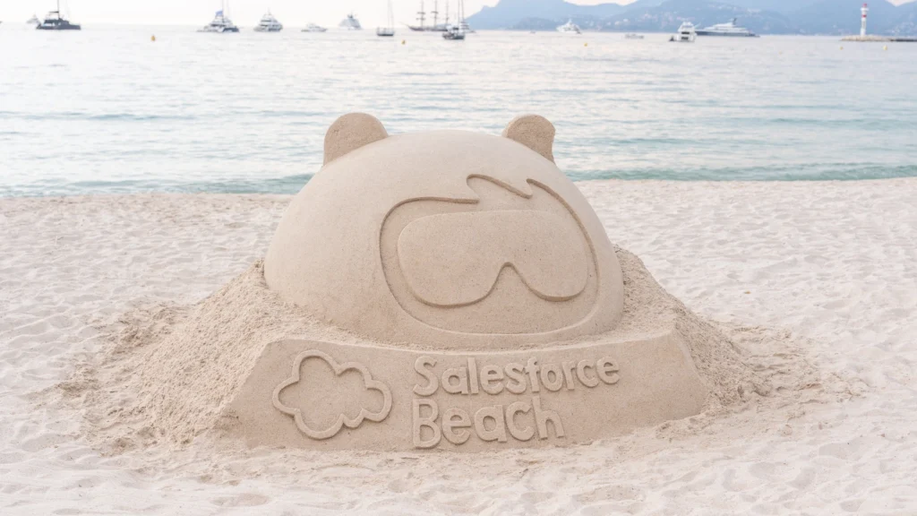 Salesforce Astro mascot sand sculpture at dusk with anchored yachts and Cannes lighthouse on the Mediterranean horizon at the Sparks-designed Salesforce Beach, Cannes Lions 2026.