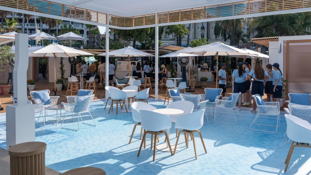 Café and bar area with light-blue tiled floor, white umbrellas, and Salesforce Beach staff in blue polos hosting attendees at the Sparks-designed Cannes Lions 2026 activation.