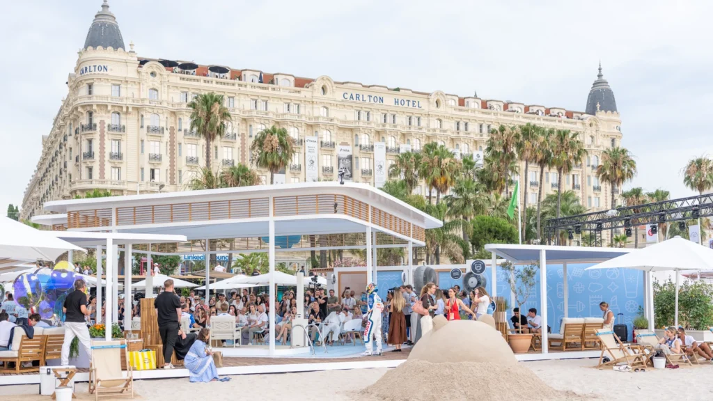 Daytime crowd around the Sparks-designed Salesforce Beach pavilion with the InterContinental Carlton Cannes hotel rising behind La Croisette at Cannes Lions 2026.