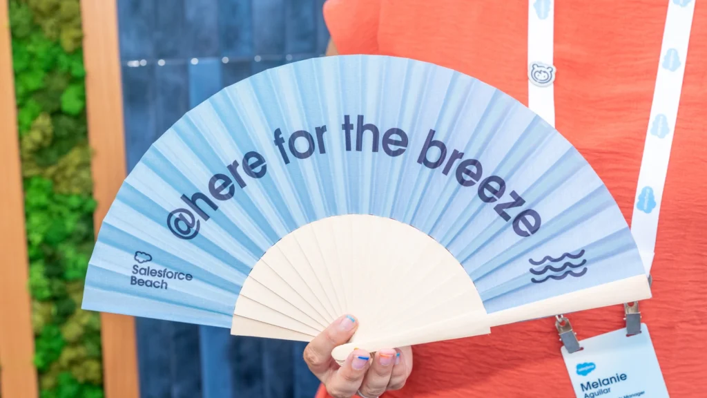 Salesforce Beach team member Melanie Aguilar holding an "@here for the breeze" branded fan against the deep blue tiled wall at the Sparks-designed Cannes Lions 2026 activation.