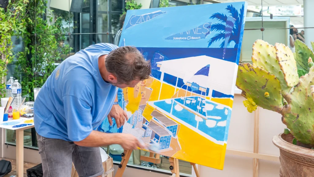 Live artist painting a stylized Salesforce Beach at Cannes canvas next to a prickly-pear cactus at the Sparks-designed Salesforce Beach activation, Cannes Lions 2026.