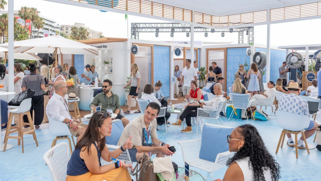 Guests seated in white lounge chairs around the Sparks-designed Salesforce Beach open-air pavilion with wood pergola, Souvenir Stop and meeting cabanas visible at Cannes Lions 2026.