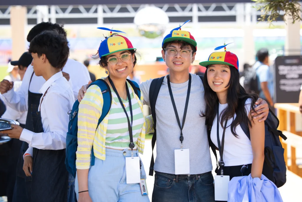 Three smiling attendees wearing Google propeller hats and I/O conference badges
