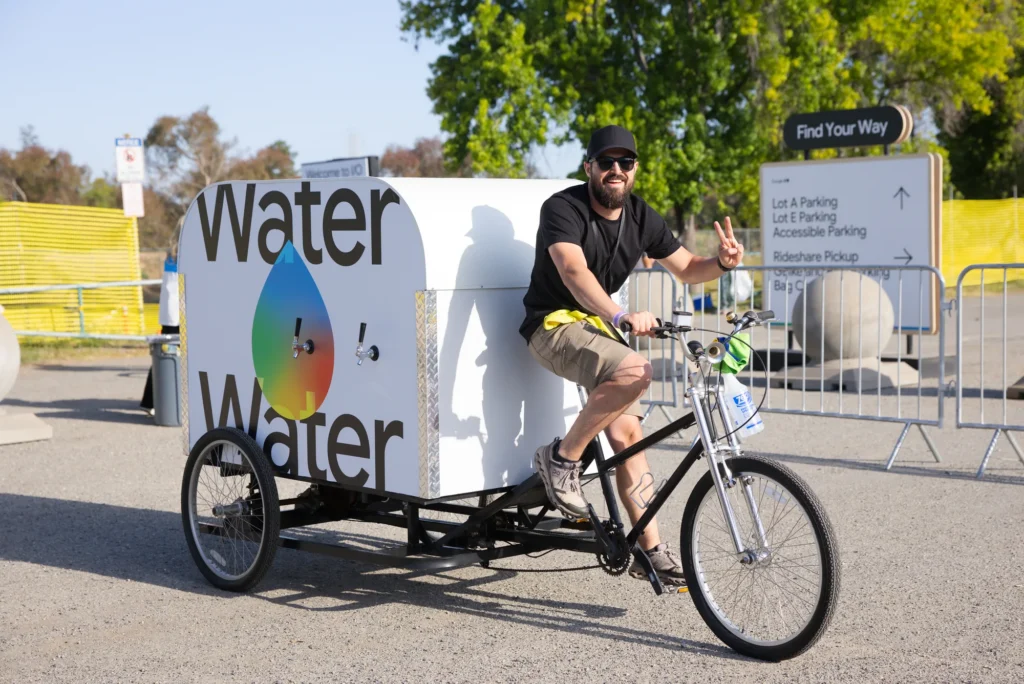 Staff member pedals a branded water-station bike across the sunny event grounds
