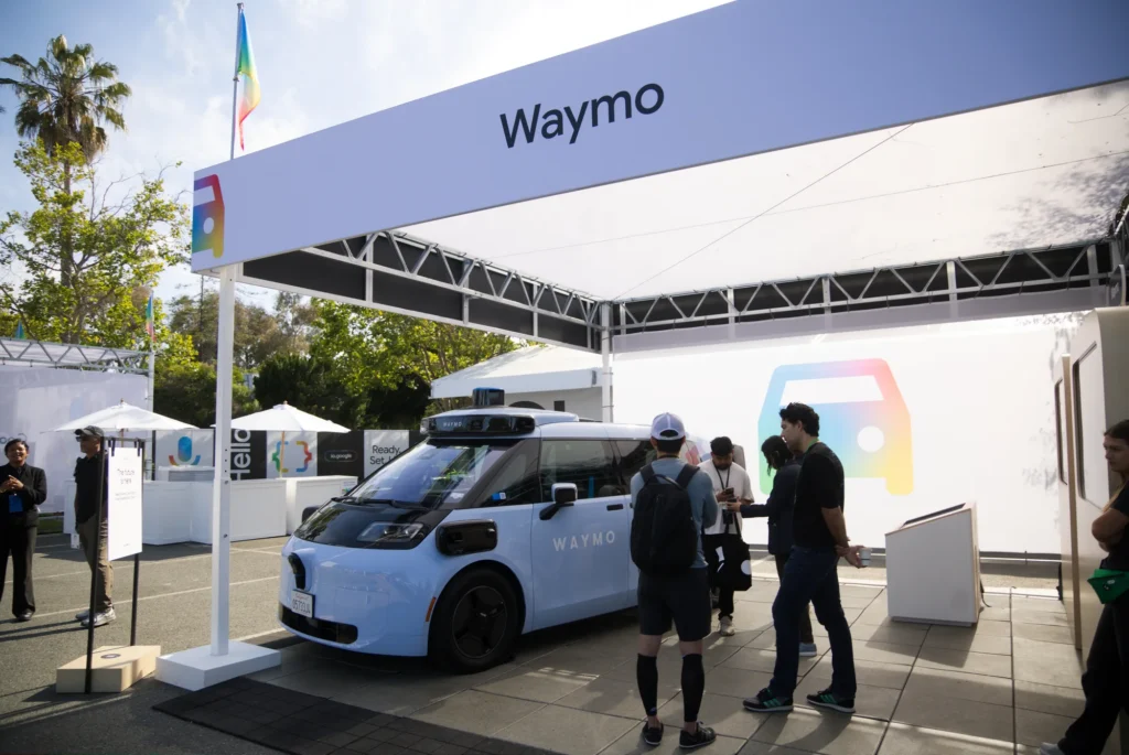 Attendees inspect a Waymo autonomous vehicle displayed under a branded canopy
