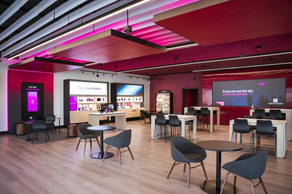 The interior of a T-Mobile retail store with magenta accent lighting, a 'one app, all the things' T Life app kiosk, Google on Android and Motorola device displays, an accessories cabinet, lounge seating, and a large screen reading 'if you can see the sky, you're connected' promoting satellite connectivity.