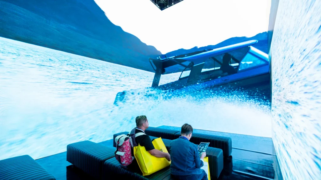 Theater screens show an electric boat racing across the water above two seated visitors