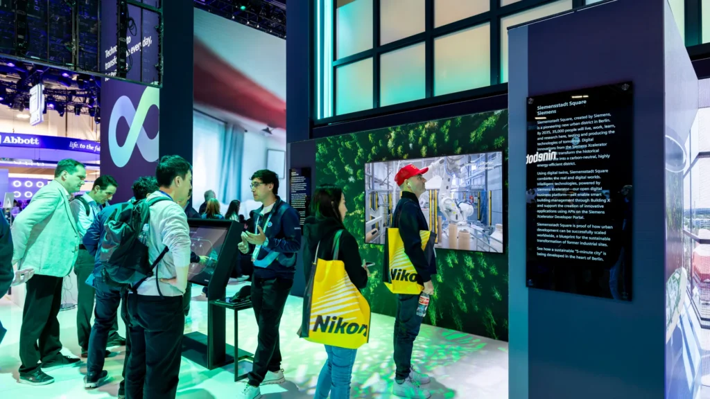 Attendees gather at a touchscreen kiosk beside a green forest video wall and plaque