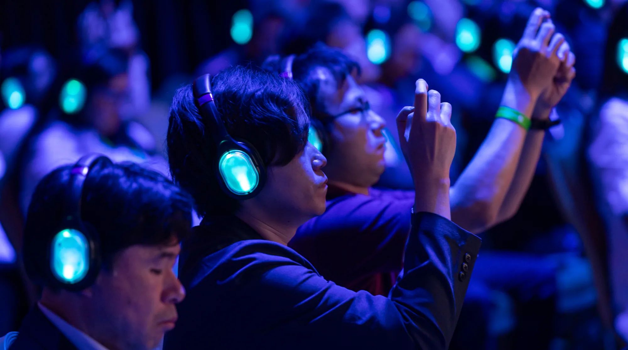 Audience members wear glowing headphones in a dimly lit room, with some raising their hands to capture the moment.
