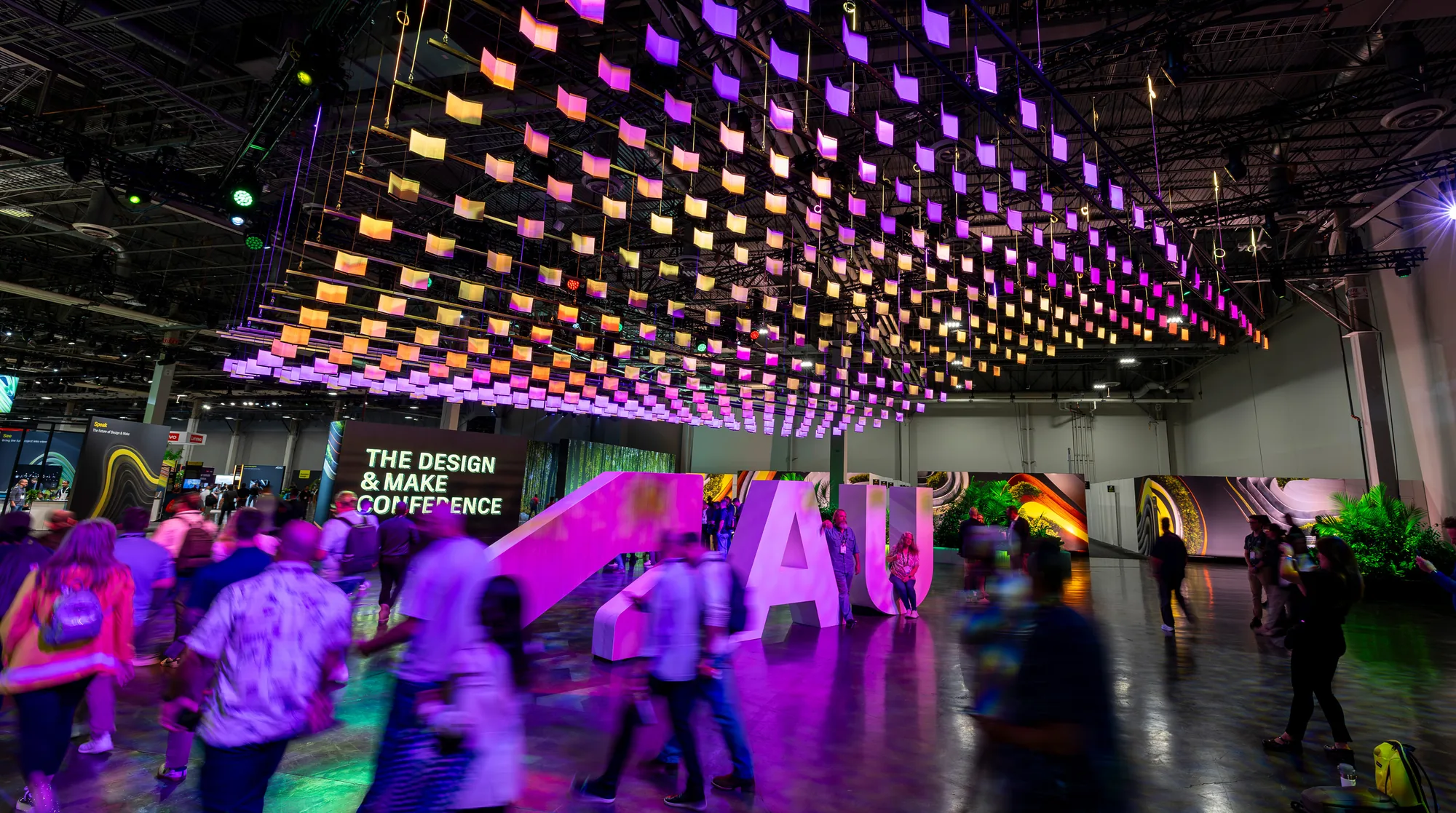 Attendees walk beneath a suspended canopy of glowing panels beside oversized AU letters at The Design & Make Conference.