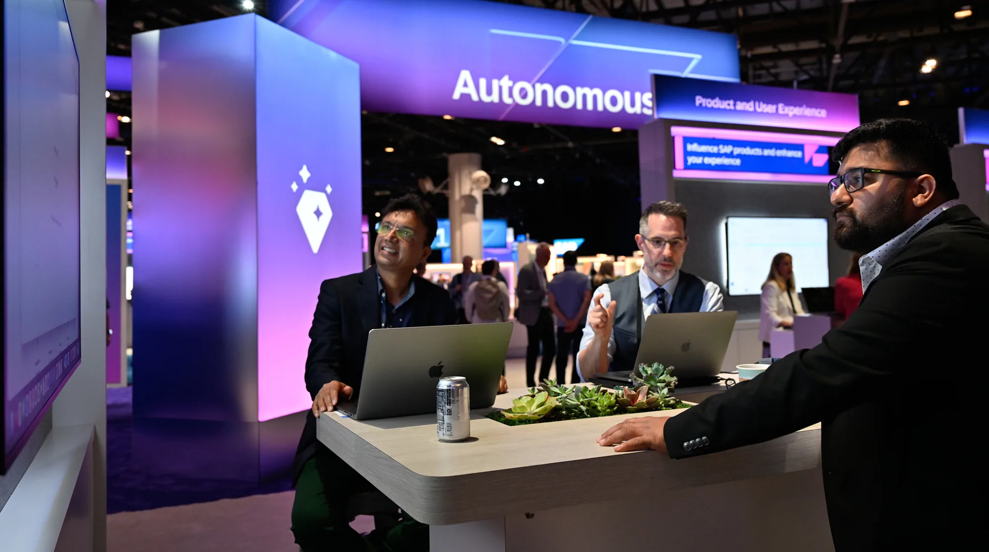 Three attendees with laptops at a high-top table in an SAP product and user experience area on the show floor.