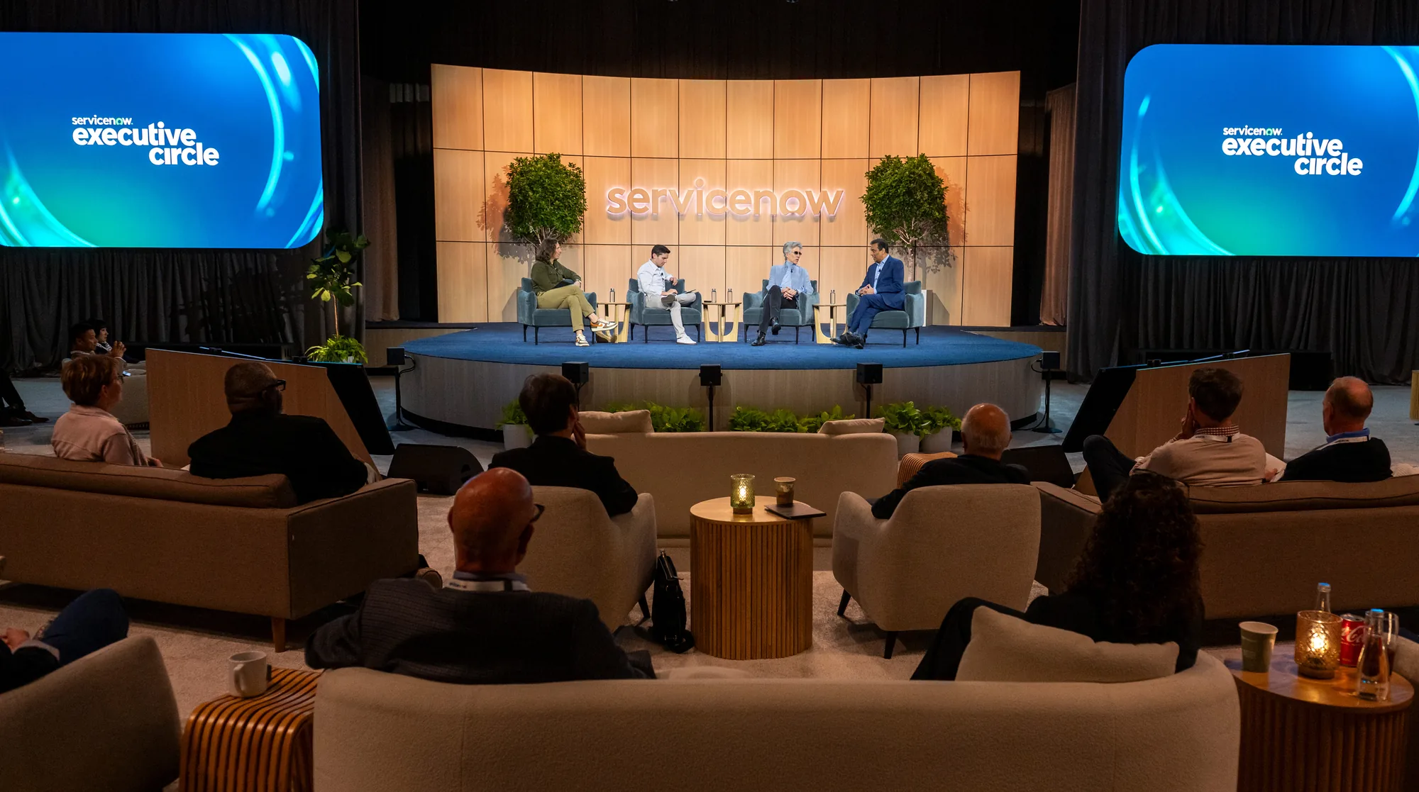 Four panelists speak on a round stage in front of a ServiceNow logo wall while guests watch from lounge seating.