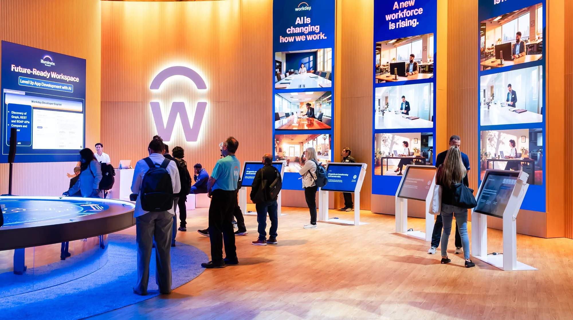 Attendees explore a Workday booth with a glowing logo wall, tall LED screens and interactive kiosks about AI.