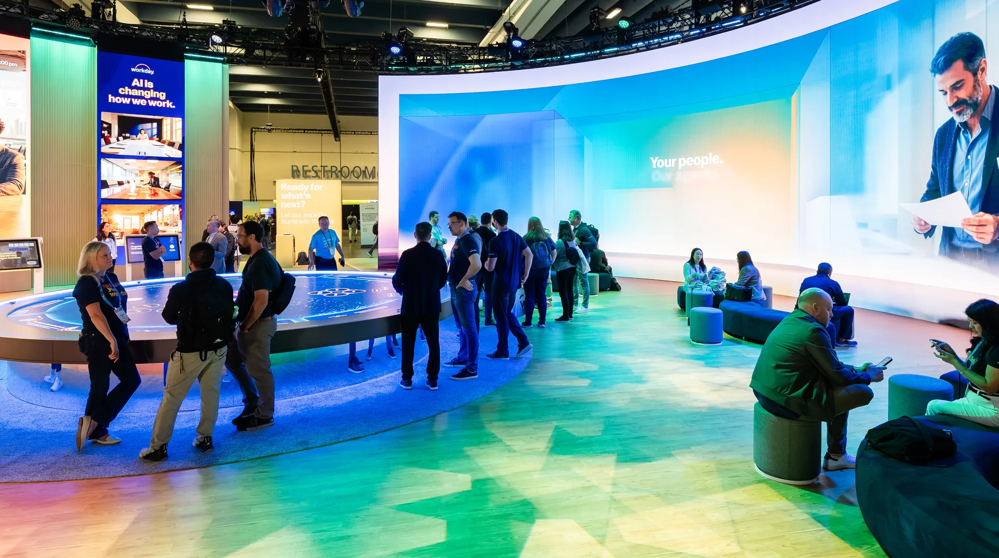 Attendees gather around a circular demo table beneath a curved LED wall reading "Your people" at a Workday booth.
